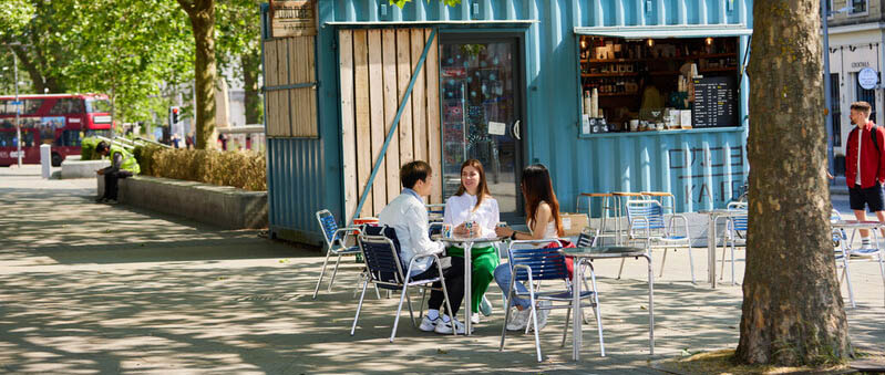 A shipping container repurposed as a coffee shop with students sat in front of it at a table